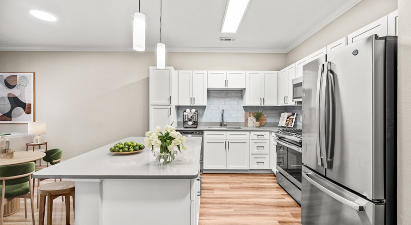 Staged kitchen at our apartments for rent  in Dedham, featuring stainless steel appliances, white cabinetry, natural wood grain plank flooring, and quartz counters with a kitchen island.